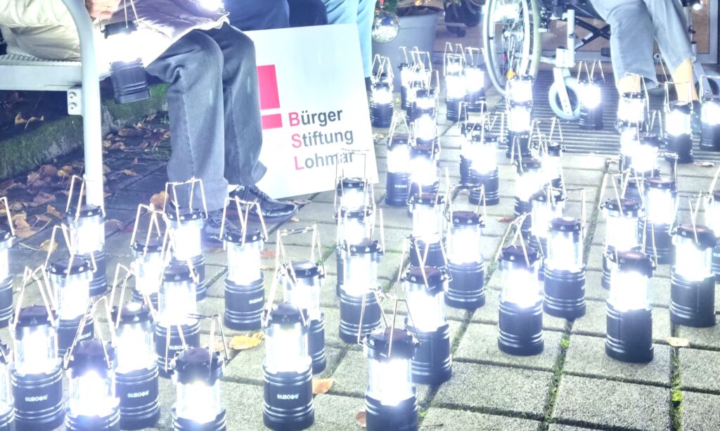 Bildausschnitt des Bildes vom Probelauf der von der Bürgerstiftung Lohmar gespendeten Tischlampe. Zu sehen sind viele leuchtende Tischlampen auf dem Boden der Terrasse vor dem Ev. Altenheim Lohmar mit dem Schild der Bürgerstiftung Lohmar.