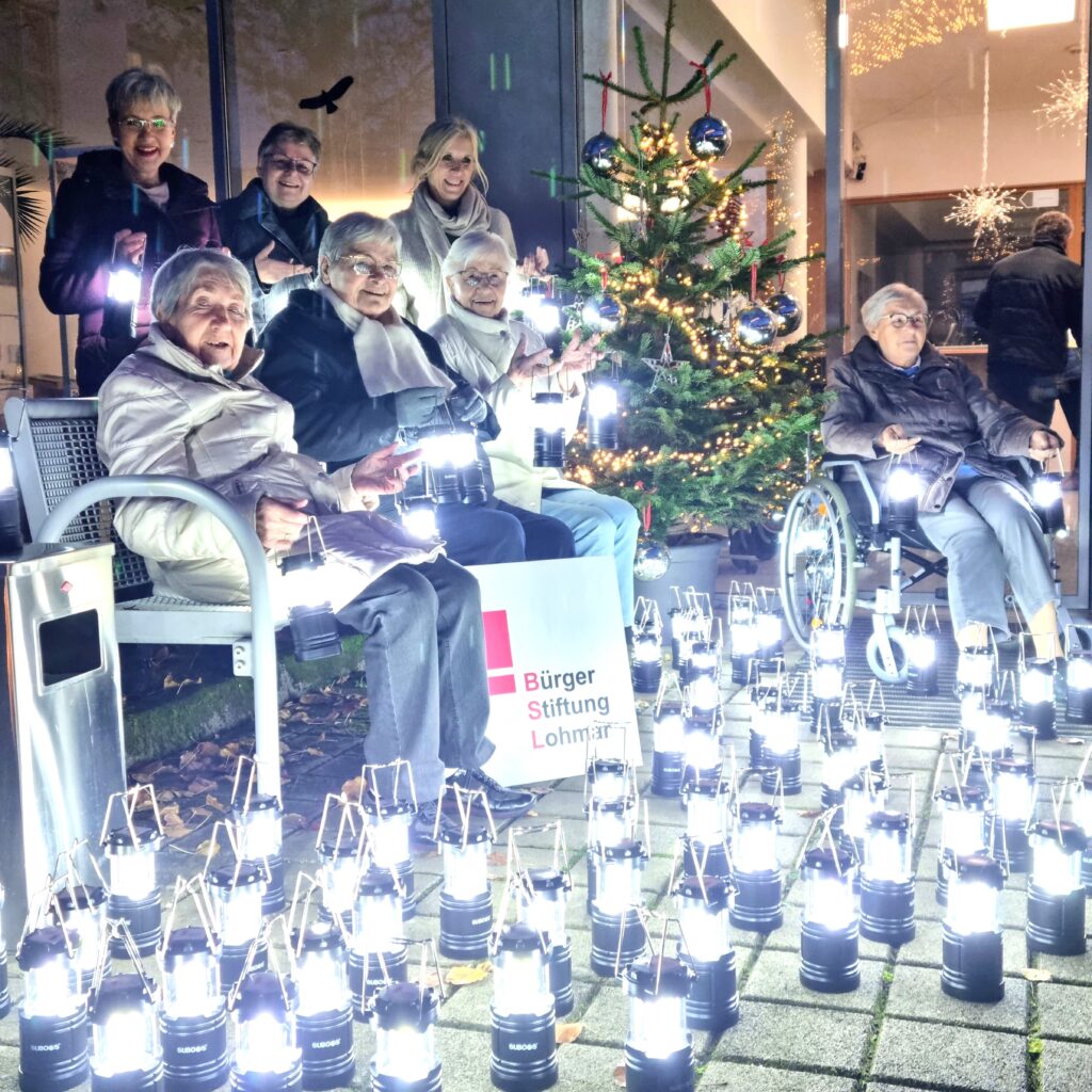 Sie strahlen mit den Lampen um die Wette (v.l.): Elisabeth Faßbender, Lieselore Neu, Anna Maria Müller und Agnes Stakemeier, dahinter Gabriele Willscheid, Yvonne Giebelen-Daughtrey und Ute Thorn-Kalsbach. Foto von sitzenden Bewohnerinnen und begleiten Personen (Namen siehe oben), die auf der Terrasse des Ev. Altenheim Lohmar hinter jede Menge leuchtender Tischlampen sitzen. Im Hintergrund ist ein geschmückter Weihnachtsbaum zu sehen.